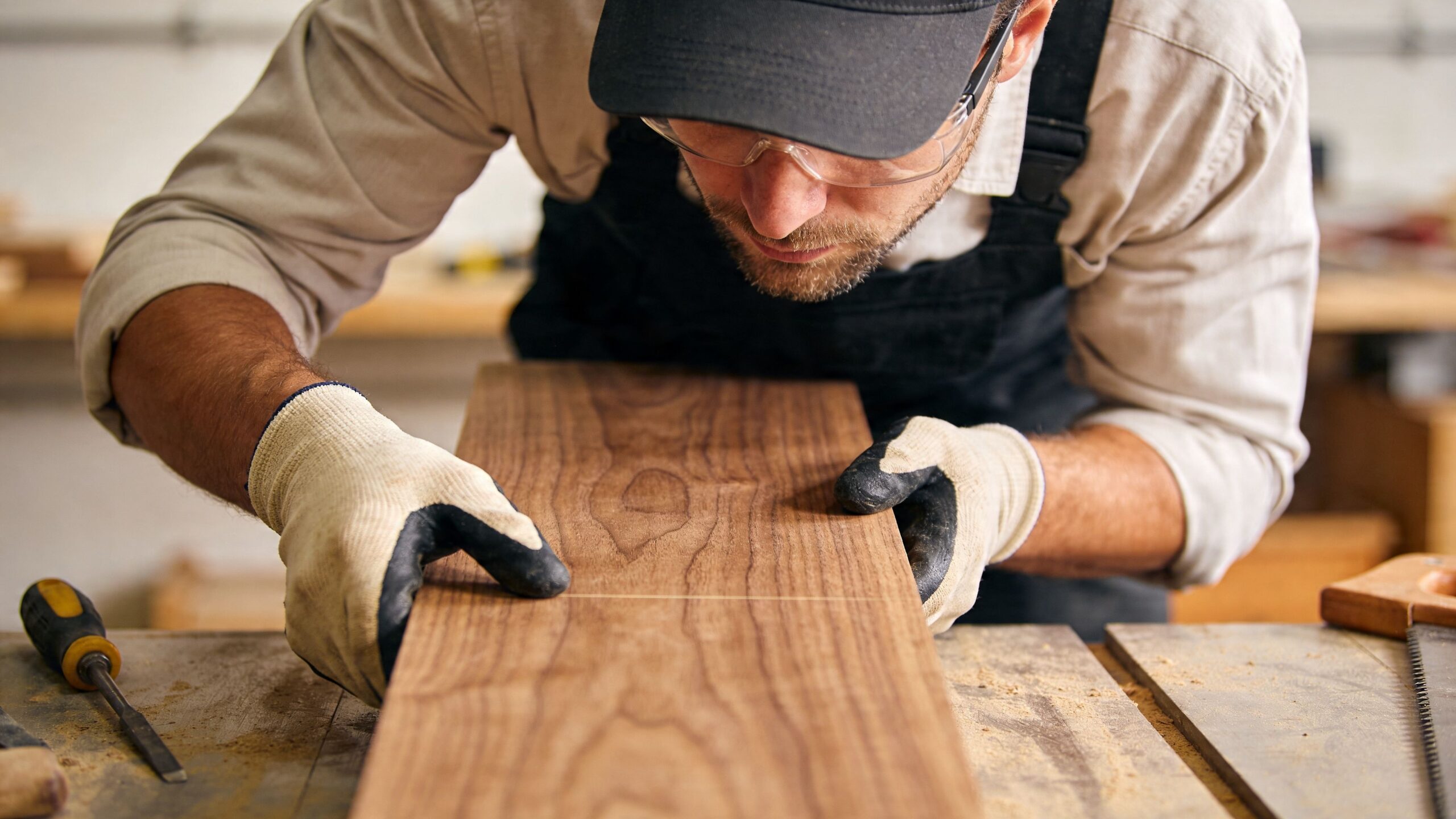 En snedker i arbejdshandsker og beskyttelsesbriller inspicerer nøje en træplanke med en opmærket streg på værkstedet.
