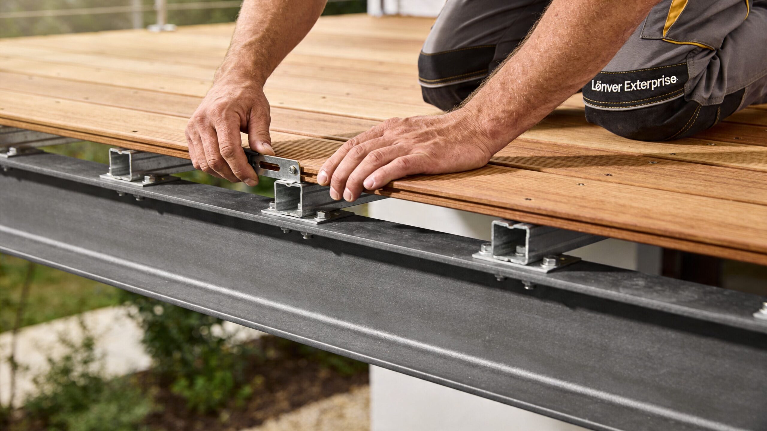 En håndværker monterer terrassebrædder af træ på en robust stålkonstruktion ved brug af et specielt monteringsbeslag.